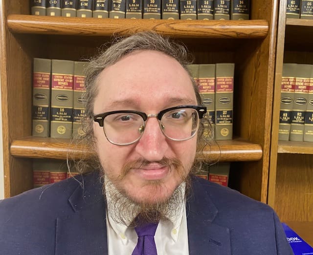 Man in suit with glasses in front of bookshelves.